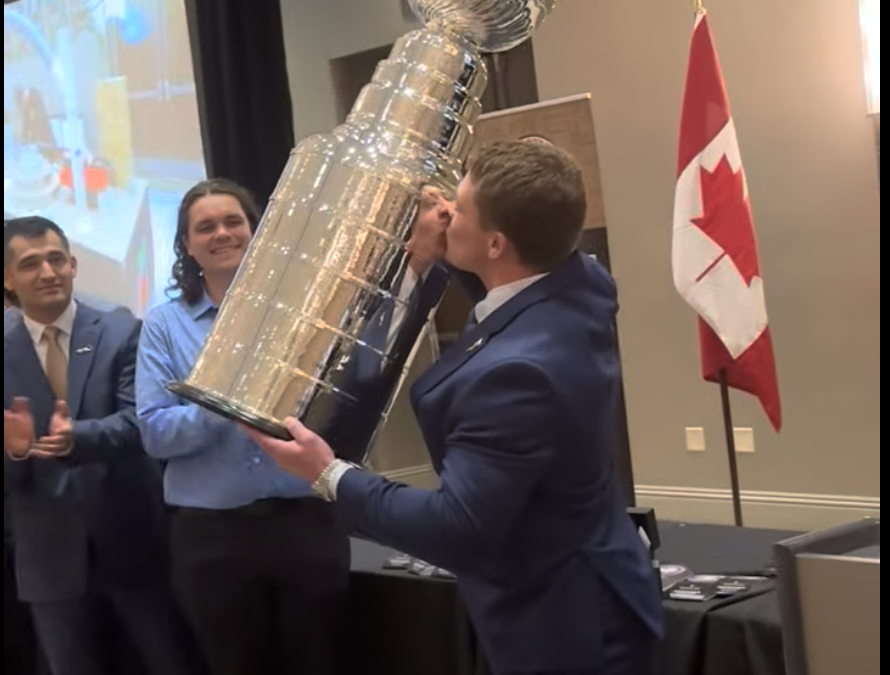 A man in a suit kisses a large trophy resembling the Stanley Cup while being applauded by others at an event. A Canadian flag is displayed in the background.